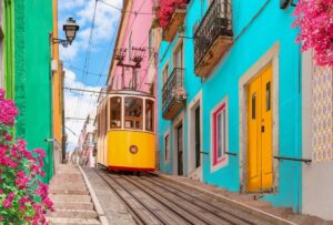 Yellow electric tram on a street with colorful houses and flowers on the balconies - Bica Elevator going down the hill of Chiado in Lisbon, Portugal.