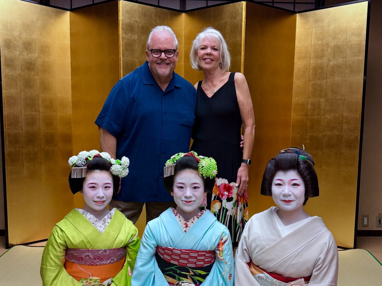David and Carol with three traditionally dressed geiko and maiko during a private cultural experience in Kyoto, Japan, posed in front of a golden folding screen.